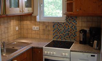 A small, modern kitchen with wooden cabinets and a bright work surface. The stove and sink are well placed next to a window with a view outside.