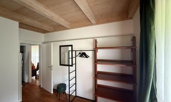 A bright room with a wooden ceiling and a large window. There is an open clothing rack and a shelf.