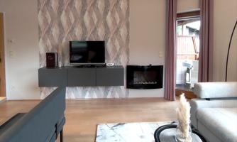 A modern living room with a TV on the wall and an elegant fireplace. The furnishings consist of a gray sofa, a table, and decorative elements.