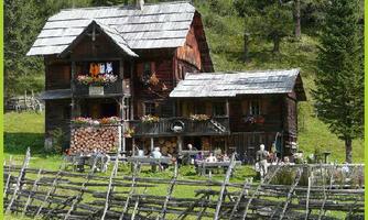 Ein malerisches Holzhaus in den Bergen mit bunten Blumen an den Fenstern. Vor dem Haus sitzen Menschen in einer einladenden Umgebung.