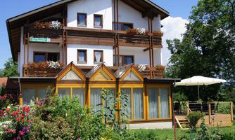 A beautiful house with blooming plants in the foreground. The terrace is equipped with a sun umbrella and the sky is blue.