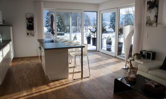 A modern kitchen with large windows and a view of a snow-covered landscape. The room is bright and friendly with wooden flooring and stylish furnishings.