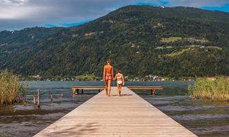 A woman and a child walk along a pier by the water. In the background, green hills and a cloudy sky can be seen.