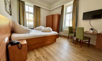 A simple room with a bed, a desk, and a television. Large windows let in plenty of daylight.