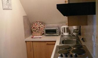 A small kitchen with a hot plate, a microwave, and a kettle. The walls are light-colored, and there are wooden cabinets as well as a decorative coffee pot.