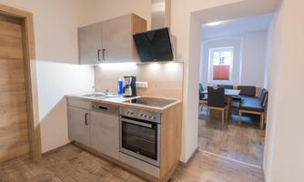A modern kitchen unit with wooden fronts and stainless steel appliances. In the back area, a dining room with a table and chairs is visible.