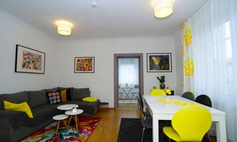 A modern living room with a cozy gray sofa and a large dining table. The walls are decorated with colorful pictures and the light is bright and friendly.