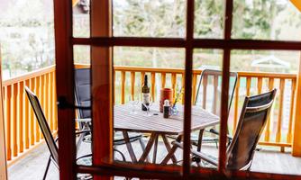 A cozy balcony with a wooden table and chairs. On the table are wine bottles and glasses, surrounded by green nature.