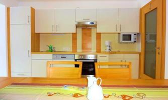 A modern, bright kitchen with wooden cabinets and a dining area. A white kettle and a colorful tablecloth are on the table.