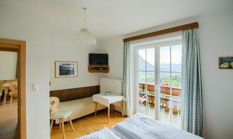 A bright room with a double bed and a small table. Large windows offer a view of nature and a balcony.