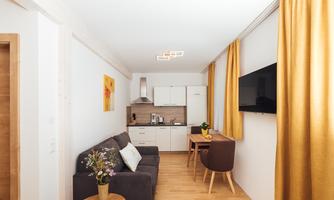 A modern apartment with a cozy sofa and a small kitchenette. The walls are bright and the curtains are in a warm yellow tone.