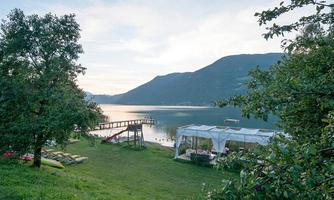 A picturesque view of a tranquil lake surrounded by mountains. In the foreground, there are trees and a meadow with a dock leading into the water.