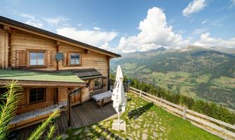 A cozy mountain cabin with wood paneling and a terrace. In the background, a beautiful green landscape stretches under a blue sky with white clouds.