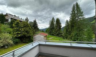 A balcony overlooking a green landscape and wooded mountains. The sky is cloudy and there are some houses nearby.