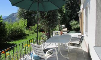A cozy outdoor area with a table and chairs under a green umbrella. In the background, a beautiful garden with trees and flowers can be seen.