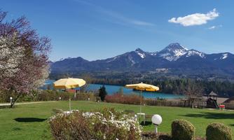 A picturesque landscape with green meadows, blooming trees, and snow-capped mountains in the background. Yellow umbrellas stand in front of a clear blue lake.