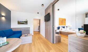 A modern hotel room with a living room and a bedroom. The decor is bright and friendly, with wooden flooring and stylish furniture.