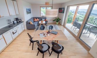 A modern living room with a kitchen and a dining area. Large windows offer a beautiful view of the surroundings.