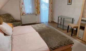 A cozy bedroom with a double bed, an armchair, and a small table. The room has large windows with curtains and is bright and cheerful in decor.