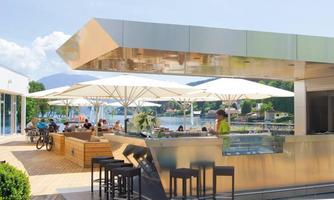 A modern bar by the water with umbrellas and many guests. The surroundings are bright and inviting, perfect for relaxed hours.