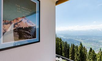 A view from a window of a picturesque mountain landscape with green trees and a wide valley view. In the foreground, a framed picture of mountains hangs on the wall.