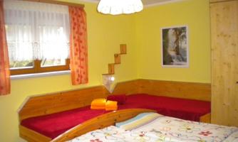 A cozy bedroom with yellow walls and two beds. The room is bright and friendly, furnished with colorful bed covers and a window with curtains.
