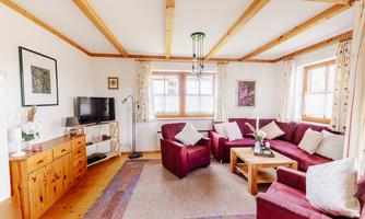 A cozy living room with red sofas and a wooden table. Large windows let in plenty of light, and the walls are decorated with pictures.