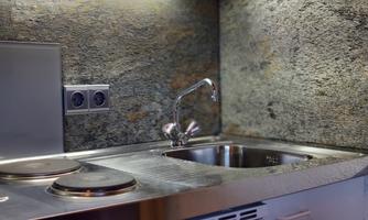 A modern kitchen corner with a stainless steel sink and two cooking plates. The walls are adorned with shimmering stone.