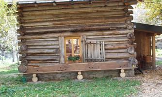 A rustic wooden house with a small window opening and a closed wooden shutter. The surroundings are lined with green grass and trees.