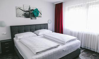 A modern bedroom with a double bed and white bed linens. Large windows let in light and there are red patterned curtains.