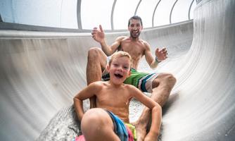 A man and a boy are laughing as they slide down a water slide. Both are swimming and having a lot of fun in the water.