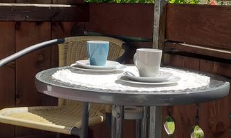 A cozy balcony with a small table and two cups. In the background, flowers and green trees are visible.