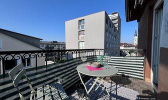 A sunny balcony with a round table and two chairs. In the background, modern buildings and a church tower can be seen.
