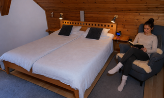 A cozy bedroom with two beds and wooden paneling. A woman is sitting comfortably in an armchair and reading a book.