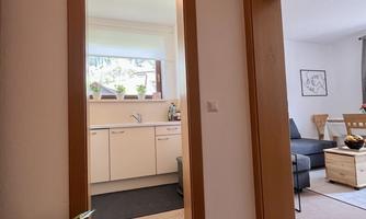 A bright hallway with a view of the kitchen and a living room in the background. The decor is modern and inviting, with plants and a rug.