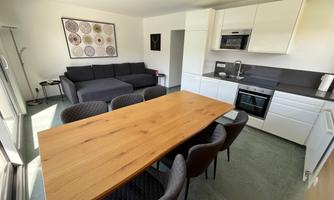 A modern kitchen with a large wooden dining table and elegant chairs. In the background, there is a cozy sofa and bright walls.