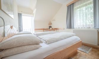A bright bedroom with a double bed and cozy pillows. The windows let in plenty of daylight, and the decor is simple and modern.