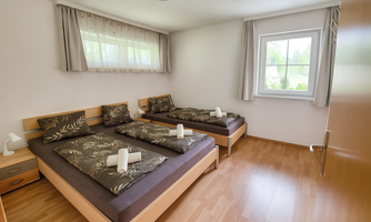 A simple bedroom with two single beds and light curtains. The room has a large window with a view outside and is well-lit.