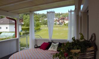 A cozy porch with a large bed and colorful cushions. In the background, green meadows and houses can be seen.