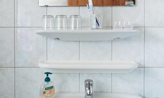 A bathroom with white tiles and a sink. On the wall, there are two shelves with glasses and care products.