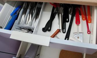 An open kitchen drawer with various cooking utensils. These include cutlery, scissors, spoons, and a cork coaster.