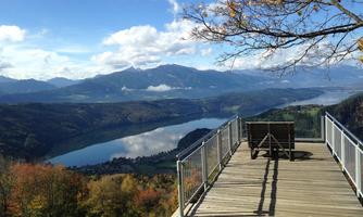 A viewing platform overlooking a clear lake and surrounding mountains. The sky is blue with some clouds, and the landscape displays autumn colors.