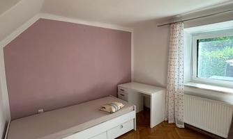 A simple bedroom with a single bed and a desk. The walls are in a soft pink, and the light comes from a window.