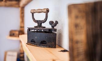 An antique iron with a chicken ornament stands on a wooden shelf. The background features a rustic wall and additional wooden details.