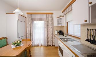 A bright kitchen with a wooden table and chairs. Large windows with curtains provide natural light.