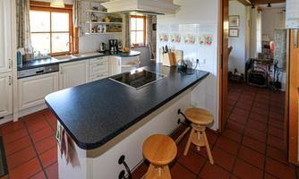 A modern kitchen with a cooking island and wooden chairs. There are windows that let in a lot of natural light, creating a cozy atmosphere.