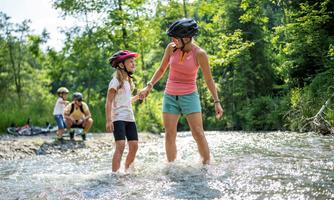 A mother and her daughter are splashing in the water of a stream. In the background, other children and bicycles can be seen.