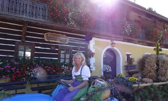 A woman in traditional attire sits on an old tractor in front of a charming farmhouse. The house is decorated with colorful flowers, and the sun is shining brightly.