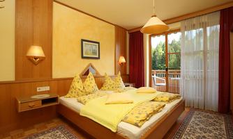 A cozy bedroom with a double bed and yellow pillows. Large windows offer a view of a balcony and plenty of natural light.