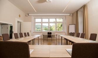 A bright meeting room with a large table and chairs. Large windows offer a view of the nature outside.